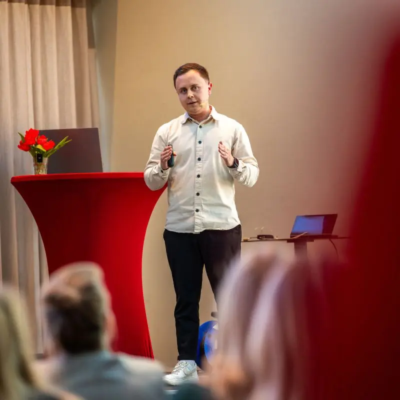 Michael Hein präsentiert auf der Bühne beim ehorses-Event „Aufsatteln im Online Marketing 2026“. Er trägt ein helles Hemd, eine dunkle Hose sowie weiße Sneaker und hält einen Presenter in der Hand. Im Vordergrund ist unscharf das Publikum zu sehen, links steht ein roter Stehtisch mit Blumen.